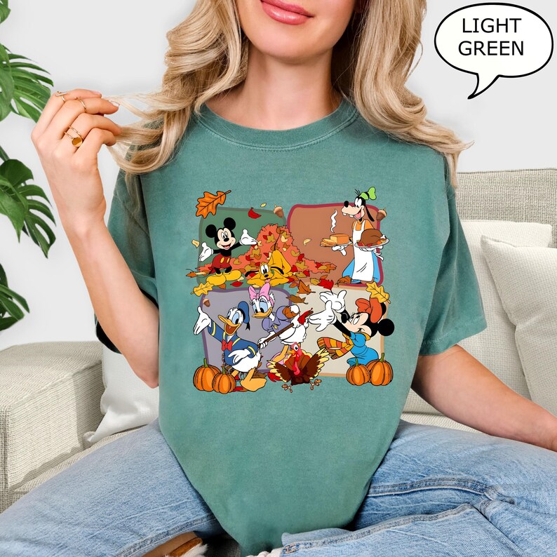 May include: Light green t-shirt featuring a colorful graphic of Disney characters celebrating Thanksgiving. The design includes Mickey Mouse, Minnie Mouse, Donald Duck, Daisy Duck, and Goofy, with pumpkins and autumn leaves. The text "LIGHT GREEN" is in a speech bubble.