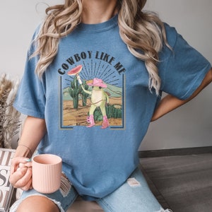 May include: A blue t-shirt with a graphic of a frog wearing a cowboy hat and holding a watermelon slice. The text "COWBOY LIKE ME" is printed on the shirt.
