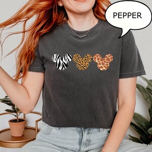 Pu&ograve; includere: T-shirt grigio scuro con tre forme di testa di Topolino con disegni animalier: zebra, leopardo e giraffa. La parola "PEPPER" &egrave; in una nuvoletta sopra la maglietta.