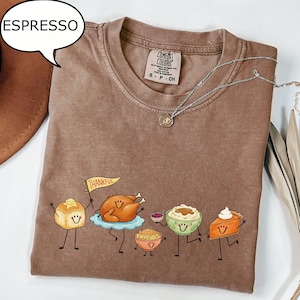 Pu&ograve; includere: Una t-shirt marrone a girocollo con una grafica di cibi del Ringraziamento antropomorfi, tra cui un panino che tiene una bandierina con la scritta THANKFUL, un tacchino arrosto, una ciotola di ripieno, pur&egrave; di patate e una fetta di torta di zucca.