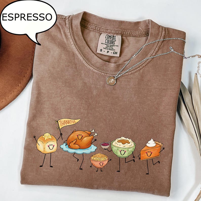 Pu&ograve; includere: Una t-shirt marrone a girocollo con una grafica di cibi del Ringraziamento antropomorfi, tra cui un panino che tiene una bandierina con la scritta THANKFUL, un tacchino arrosto, una ciotola di ripieno, pur&egrave; di patate e una fetta di torta di zucca.