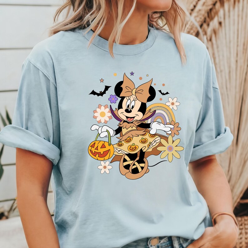 Puede incluir: Camiseta azul claro con un dise&ntilde;o de Minnie Mouse con tem&aacute;tica de Halloween. Minnie Mouse est&aacute; disfrazada, sosteniendo una linterna de calabaza, con murci&eacute;lagos, flores y un arco&iacute;ris. La camiseta tiene mangas enrolladas.