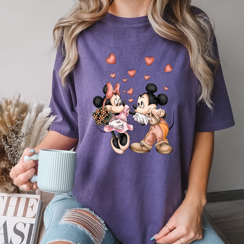 May include: Purple t-shirt with a graphic of Mickey Mouse and Minnie Mouse holding hands with hearts around them.