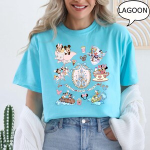 Puede incluir: Camiseta azul claro con varios personajes de dibujos animados y el texto "Best Day Ever". La camiseta tiene cuello redondo y mangas cortas. El gr&aacute;fico incluye im&aacute;genes de un castillo, tazas de t&eacute; y otros elementos tem&aacute;ticos.