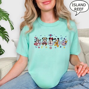 May include: A light teal t-shirt featuring a nautical-themed design with cartoon characters in circular frames. The design includes the text "ISLAND REEF" in a speech bubble. The characters are dressed in sailor outfits.