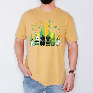 Puede incluir: Camiseta de color amarillo mostaza con un gr&aacute;fico de dibujos animados de personajes de Star Wars frente a una silueta de castillo. Los personajes incluyen a R2-D2, un Stormtrooper, Darth Vader, Boba Fett y otros. Tr&eacute;boles verdes y globos amarillos est&aacute;n encima del castillo.