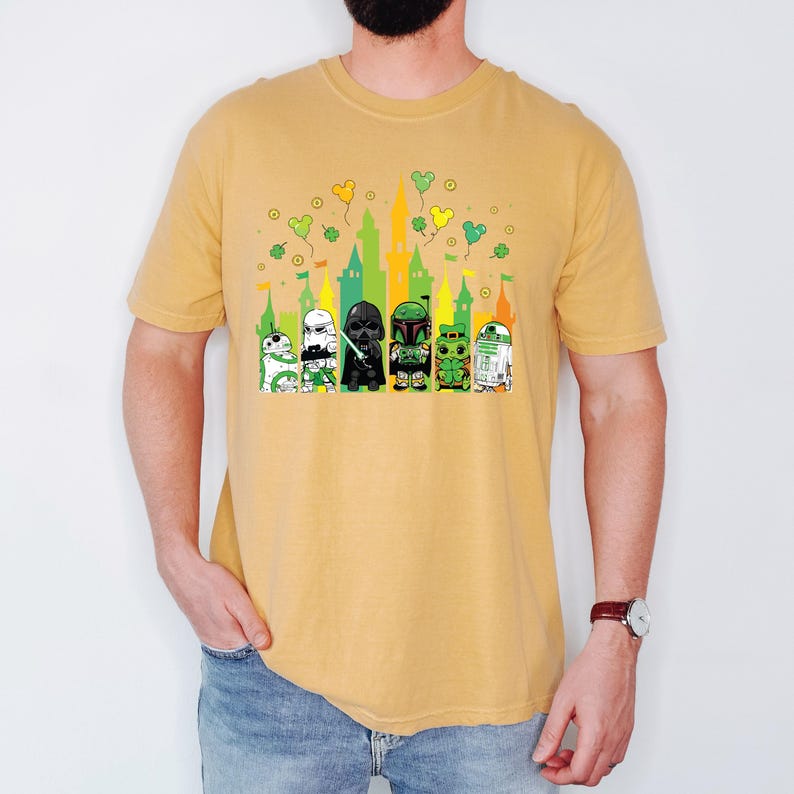 Puede incluir: Camiseta de color amarillo mostaza con un gr&aacute;fico de dibujos animados de personajes de Star Wars frente a una silueta de castillo. Los personajes incluyen a R2-D2, un Stormtrooper, Darth Vader, Boba Fett y otros. Tr&eacute;boles verdes y globos amarillos est&aacute;n encima del castillo.