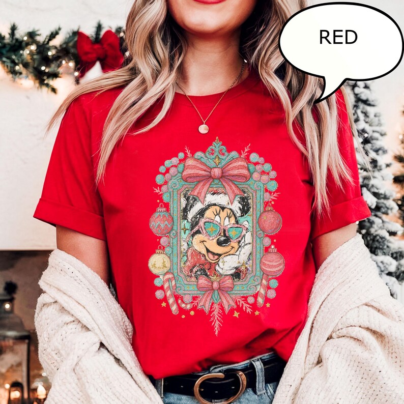Puede incluir: Camiseta roja con un dise&ntilde;o de Minnie Mouse con gafas de sol y gorro de Pap&aacute; Noel, rodeada de adornos navide&ntilde;os. La palabra "RED" est&aacute; en un bocadillo.