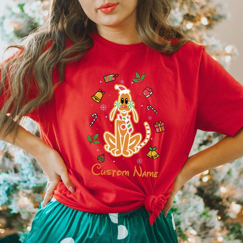 May include: Red t-shirt with a gingerbread dog wearing a Santa hat and surrounded by Christmas decorations. The text "Custom Name" is printed below the dog.