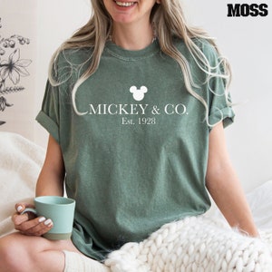 Puede incluir: Una camiseta verde con una cabeza de Mickey Mouse blanca y el texto "MICKEY & CO. Est. 1928"
