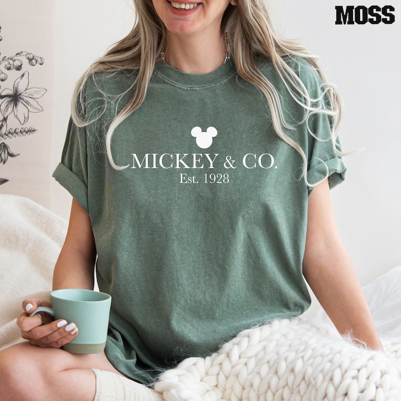 Puede incluir: Una camiseta verde con una cabeza de Mickey Mouse blanca y el texto "MICKEY & CO. Est. 1928"