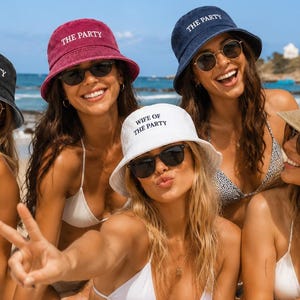 May include: A collection of five bucket hats in black, magenta, navy blue, white, and tan. Each hat features white embroidered text on the front. Four hats display the phrase THE PARTY, while the white hat displays WIFE OF THE PARTY.