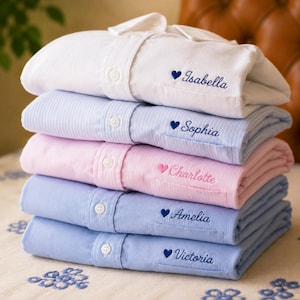 May include: A stack of five folded long-sleeve button-down shirts in white, light blue pinstripe, light pink pinstripe, and two shades of blue. Each shirt features an embroidered heart and a name: Isabella, Sophia, Charlotte, Amelia, and Victoria.
