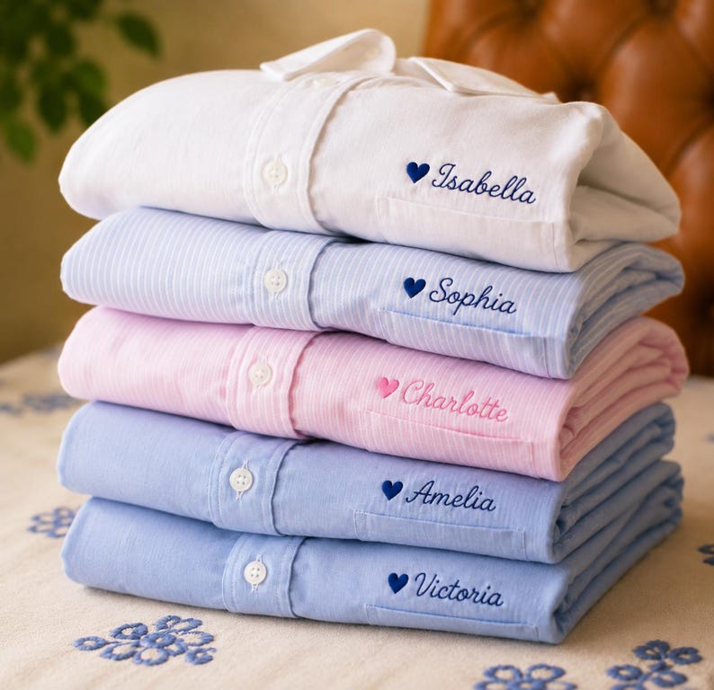 May include: A stack of five folded long-sleeve button-down shirts in white, light blue pinstripe, light pink pinstripe, and two shades of blue. Each shirt features an embroidered heart and a name: Isabella, Sophia, Charlotte, Amelia, and Victoria.