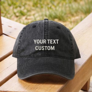 Op de afbeelding: Een zwarte baseball cap met de tekst "YOUR TEXT CUSTOM" in het wit geborduurd. De cap is gemaakt van een zacht, versleten materiaal en heeft een gebogen klep. De cap ligt op een houten oppervlak.