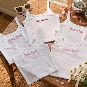 May include: White ribbed tank tops with pink embroidered text, including "Bride's Babes," "Bride Squad," and "Mrs. Gunter." The image also features heart-shaped sunglasses, disco balls, and flowers, suggesting a bridal theme.