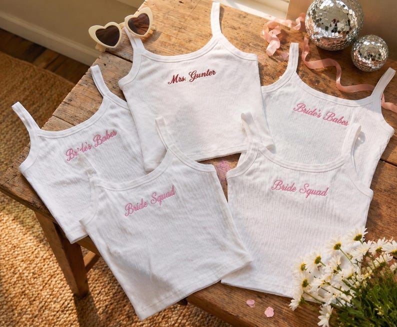 May include: White ribbed tank tops with pink embroidered text, including "Bride's Babes," "Bride Squad," and "Mrs. Gunter." The image also features heart-shaped sunglasses, disco balls, and flowers, suggesting a bridal theme.