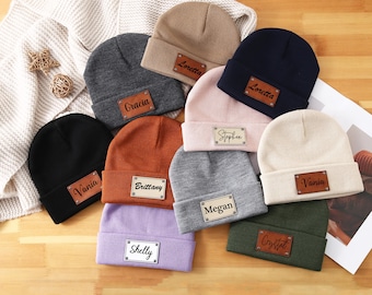May include: A collection of personalized beanies in various colors, including black, navy, and gray. Each beanie features a rectangular leather patch with a name, such as "Vania" or "Loretta". The hats are arranged on a wooden surface.