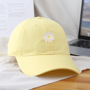 Op de afbeelding: Lichtgele baseballcap met een witte madelief geborduurd op de voorkant. De cap heeft een gebogen klep en een klassiek zes-panelen ontwerp. De hoed is gemaakt van een zachte stof en is geschikt voor casual wear.