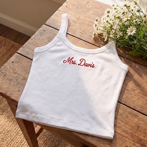 May include: White ribbed tank top with the embroidered text "Mrs. Davis" in red script. The top has a scoop neckline and thin straps. The garment is displayed on a rustic wooden surface.