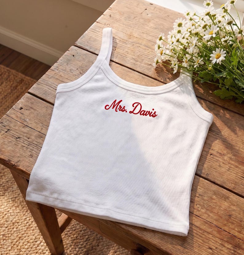 May include: White ribbed tank top with the embroidered text "Mrs. Davis" in red script. The top has a scoop neckline and thin straps. The garment is displayed on a rustic wooden surface.