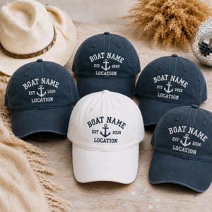 May include: A collection of five baseball caps, four in dark navy blue and one in white. Each cap features an embroidered anchor graphic with the text BOAT NAME, EST 2026, and LOCATION. The caps have a curved brim and a structured crown design.