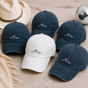 Könnte beinhalten: Vier marineblaue Baseballkappen und eine weiße Baseballkappe, jede mit einem kleinen, weiß gestickten Wellenmotiv auf der Vorderseite. Die Kappen verfügen über eine strukturierte Krone, einen gebogenen Schirm und einen verstellbaren Riemen.