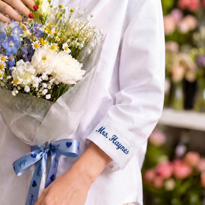May include: A white long-sleeved shirt with the name Mrs. Haynes embroidered in blue on the cuff. The shirt is paired with a bouquet of white, blue, and yellow flowers wrapped in translucent paper and tied with a blue ribbon printed with hearts.