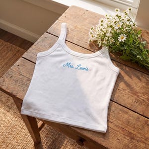 May include: White ribbed tank top with the embroidered text "Mrs. Lewis" in light blue. The top is displayed on a rustic wooden table, with a bouquet of white daisies nearby. The tank top has a scoop neckline and thin straps.