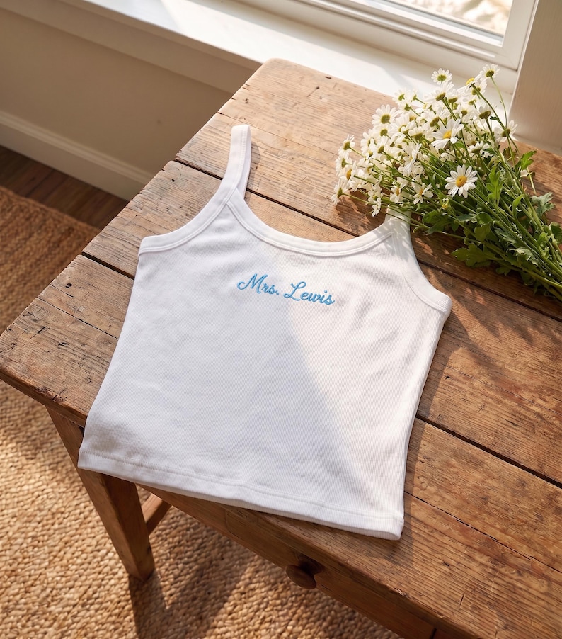 May include: White ribbed tank top with the embroidered text "Mrs. Lewis" in light blue. The top is displayed on a rustic wooden table, with a bouquet of white daisies nearby. The tank top has a scoop neckline and thin straps.