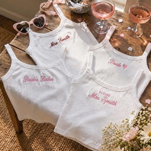 May include: Four white ribbed tank tops with pink embroidered text. One says "Mrs. Smith", another "Bride", a third "Future Mrs. Smith", and the last "Bride's Babes". Pink heart-shaped sunglasses and two glasses of pink champagne are also visible.