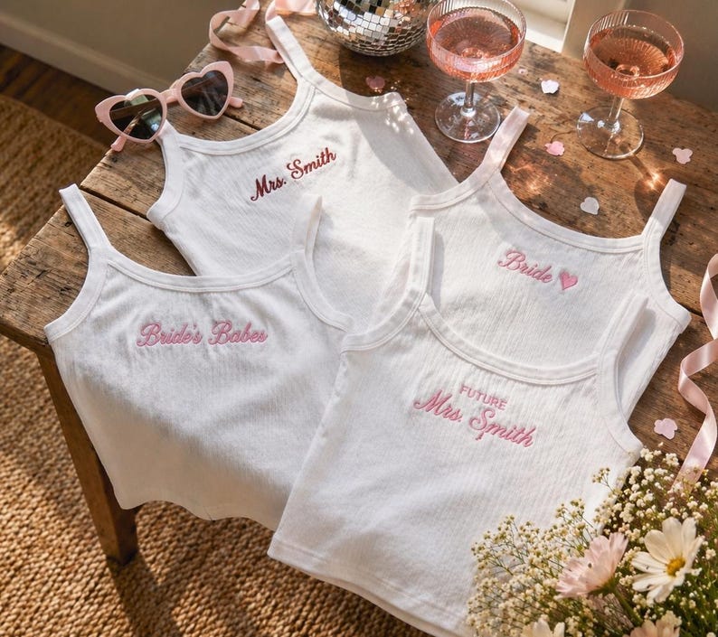 May include: Four white ribbed tank tops with pink embroidered text. One says "Mrs. Smith", another "Bride", a third "Future Mrs. Smith", and the last "Bride's Babes". Pink heart-shaped sunglasses and two glasses of pink champagne are also visible.