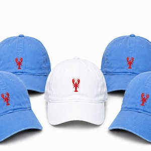 May include: Four light blue baseball caps and one white baseball cap arranged in a group. Each cap features a small, embroidered red lobster centered on the front panel. The caps have a curved brim, a structured crown, and eyelet ventilation holes.