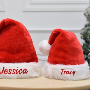 Op de afbeelding: Twee rode kerstmanmutsen met witte bontkraag. De mutsen zijn gepersonaliseerd met de namen "Jessica" en "Tracy" in rode borduurwerk.