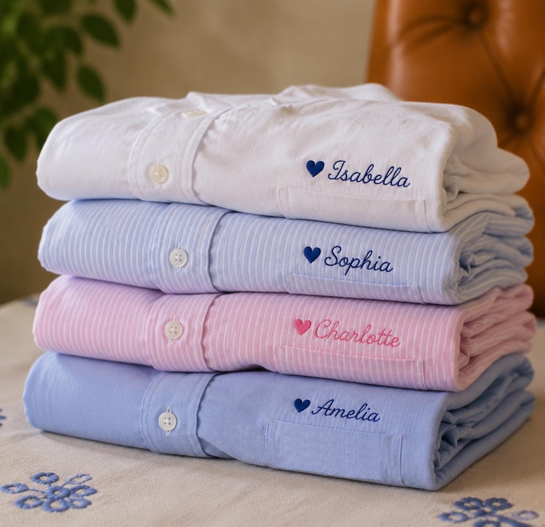 May include: A stack of four folded button-down shirts in white, light blue striped, pink striped, and solid light blue. Each shirt features a chest pocket embroidered with a small heart and a name: Isabella, Sophia, Charlotte, or Amelia.