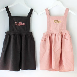 May include: Two sleeveless pinafore dresses, one brown and one pink, hanging on white hangers. The brown dress has the word "Custom" embroidered on the front in pink thread. The pink dress has the word "Elise" embroidered on the front in gold thread.
