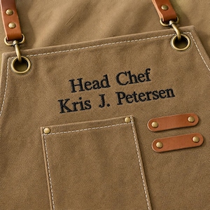 May include: A tan canvas apron featuring brown leather straps attached with brass-toned metal hardware. The apron is embroidered with the black text "Head Chef" and "Kris J. Petersen" above a front pocket with two decorative leather tabs.