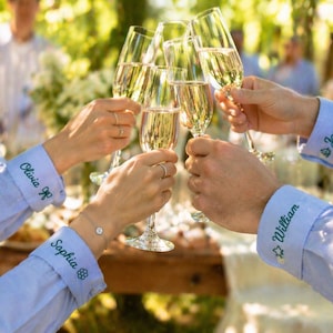 May include: Several champagne flutes are raised in a celebratory toast. The glasses are filled with a golden liquid. The sleeves of the light blue shirts have names embroidered in green: Olivia, Sophia, William, and James. The background is blurred with greenery.