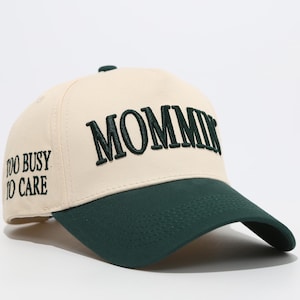 May include: Cream and dark green baseball cap. The word "MOMMIN" is embroidered in dark green across the front, with "TOO BUSY TO CARE" embroidered on the side. A fashionable accessory for casual wear.