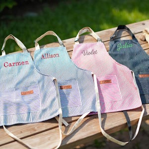 May include: Four striped aprons with names embroidered on them: Carmen, Allison, Violet, and Julia. Each apron has a pocket with a leather tag that says "Apron". The aprons are on a wooden table with gardening gloves and a small potted plant.