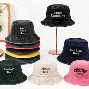 May include: A collection of bucket hats in various colors including black, navy, pink, green, and cream. Some hats feature embroidered text such as Your Logo Here, Custom Embroidered, or Custom Text. The hats have a soft, washed fabric texture.
