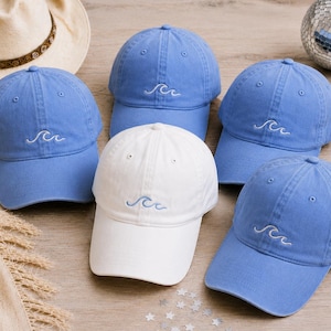 May include: A collection of five baseball caps, four in light blue and one in white, each featuring a small white embroidered wave design on the front panel. The caps have a structured crown, a curved brim, and eyelets for ventilation.