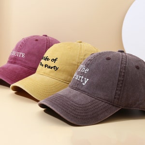 May include: Three baseball caps in a row, each with a different colour and text. The first is a deep red with "CSUITE" embroidered in white. The second is yellow with "Wife of the Party" in black. The third is brown with "The Party" in white.
