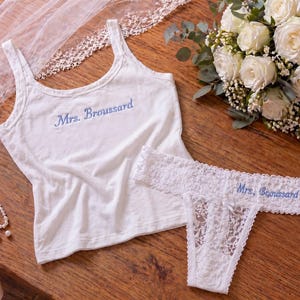 May include: A white bridal lingerie set with blue embroidered text reading "Mrs. Broussard". The set includes a tank top and a lace thong. A bouquet of white roses and a veil are also present.