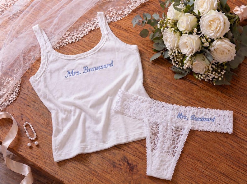 May include: A white bridal lingerie set with blue embroidered text reading "Mrs. Broussard". The set includes a tank top and a lace thong. A bouquet of white roses and a veil are also present.