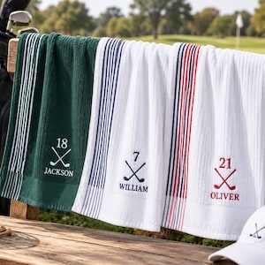 Può includere: Tre asciugamani da golf rettangolari in verde scuro e bianco, con strisce verticali e disegni ricamati. Ogni asciugamano mostra un numero sopra mazze da golf incrociate e un nome: 18 Jackson, 7 William e 21 Oliver in rosso.