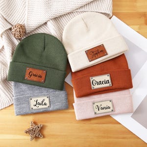 May include: Five knitted beanies with leather name tags. The beanies are in various colors, including green, beige, gray, orange, and pink. The names on the tags are Stephen, Gracia, Laila, Gracia, and Vania.