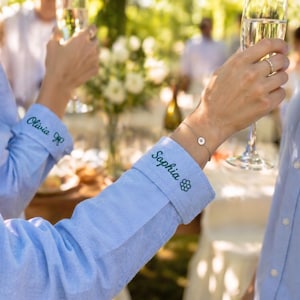 May include: Two light blue long-sleeved button-down shirts with personalized embroidery on the cuffs. One cuff features the name Olivia with a small bow icon, and the other cuff displays the name Sophia with a small flower icon in dark thread.