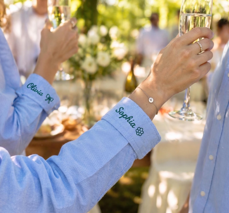May include: Two light blue long-sleeved button-down shirts with personalized embroidery on the cuffs. One cuff features the name Olivia with a small bow icon, and the other cuff displays the name Sophia with a small flower icon in dark thread.