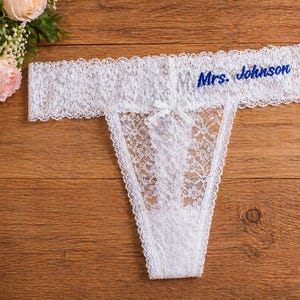 May include: White lace thong with the embroidered text "Mrs. Johnson" in blue. The lingerie has a delicate floral pattern and a small bow. The thong is displayed on a wooden surface, with a bouquet of roses in the background.